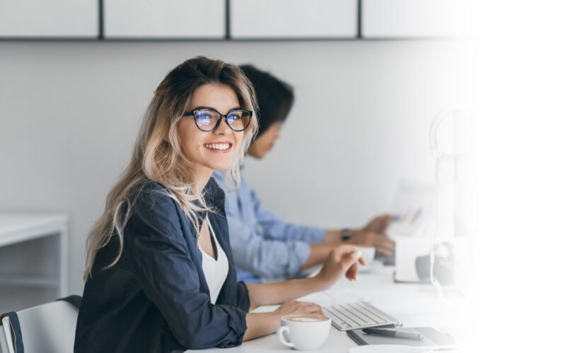 Attractive laughing freelancer girl posing with cup of coffee at her workplace. Chinese student in blue shirt works with document in campus with blonde friend in glasses.